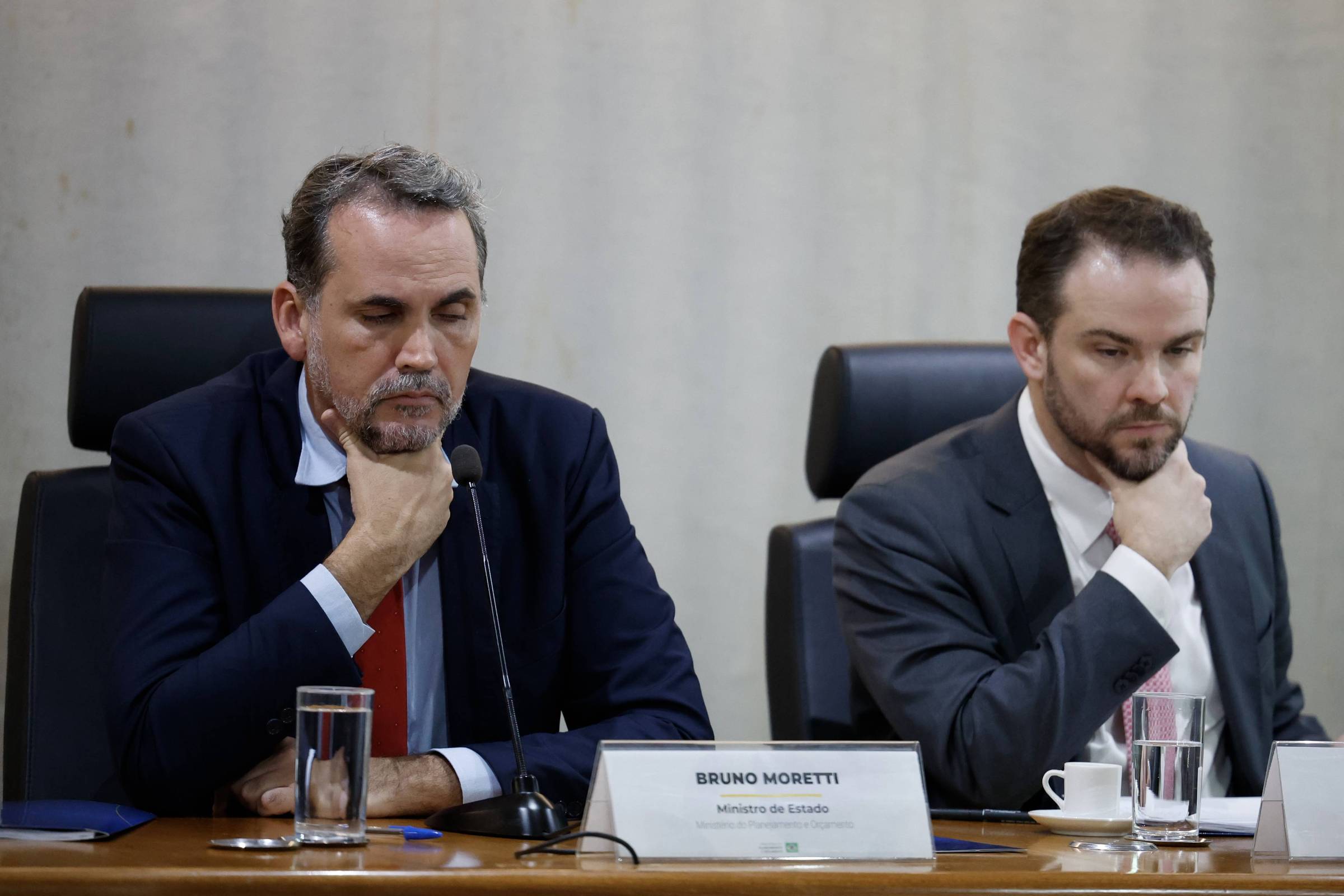 Dois homens de terno escuro sentados em cadeiras pretas em mesa de reunião. Ambos apoiam a mão no queixo, com expressão séria. À frente do homem à esquerda, placa com nome