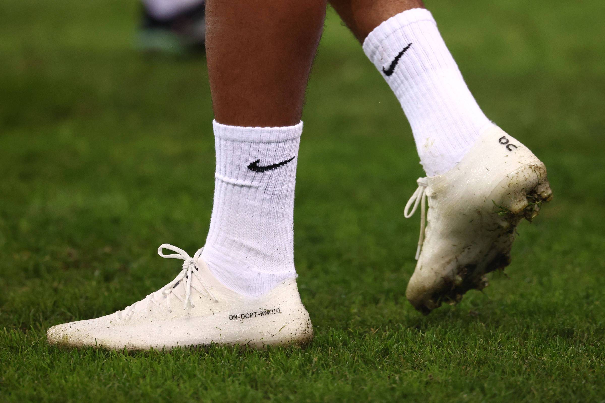 Pés de jogador com chuteiras brancas e meias brancas com logo da Nike, em campo de grama verde.