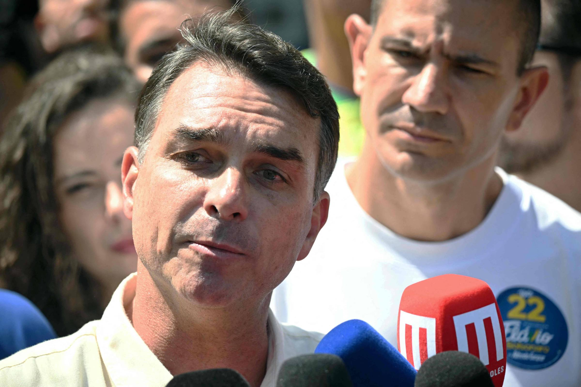 Homem de cabelo curto e grisalho fala para vários microfones em coletiva ao ar livre. Pessoas ao fundo observam atentamente, incluindo homem com camiseta branca e adesivo azul.