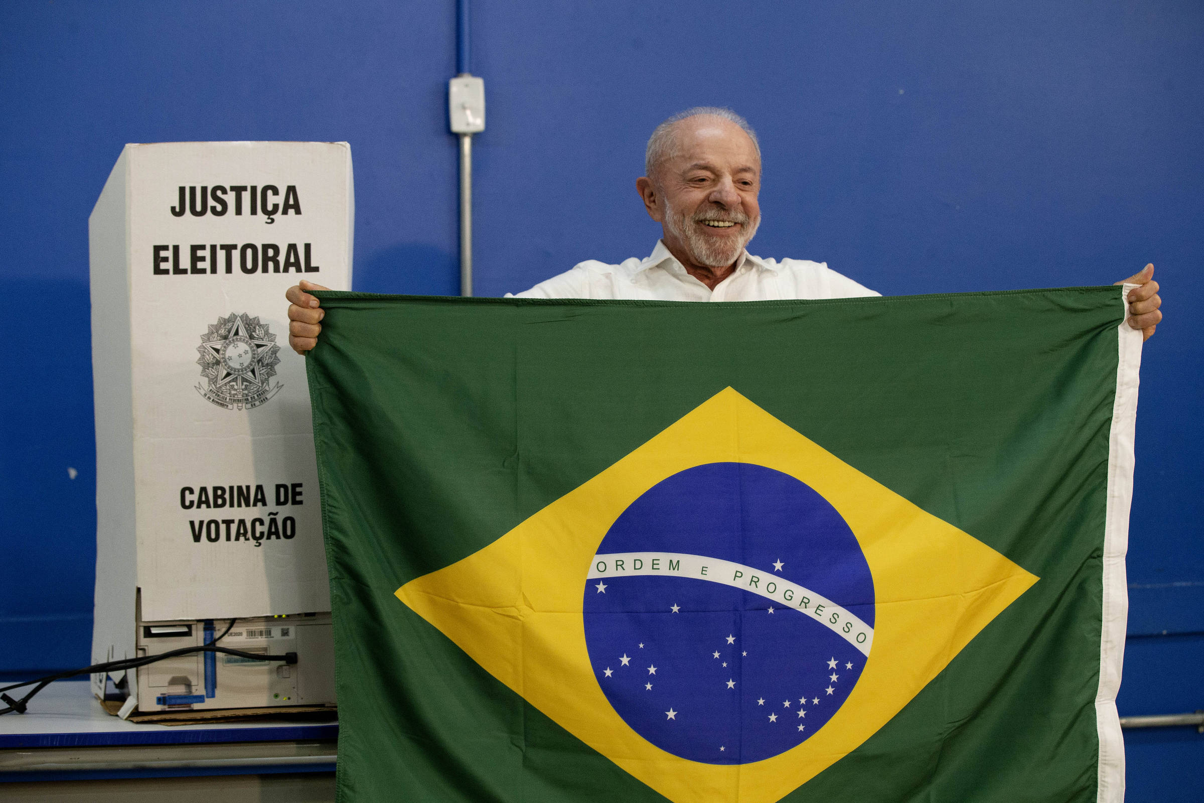 Homem sorridente segura a bandeira do Brasil aberta em frente ao corpo, em ambiente com parede azul. À esquerda, urna eletrônica com identificação