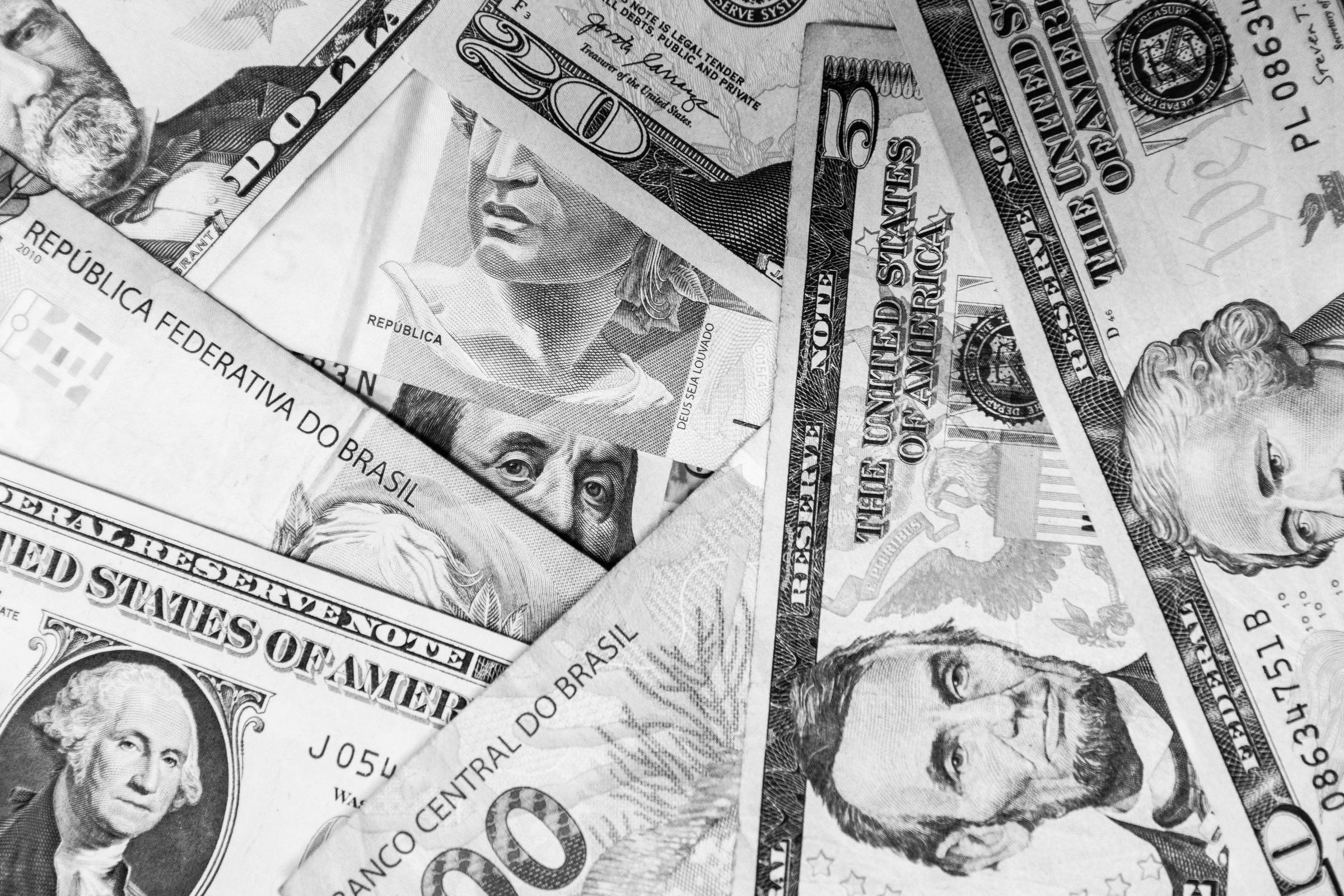 A imagem mostra uma variedade de notas de dinheiro, incluindo cédulas do Brasil e dos Estados Unidos. As cédulas estão dispostas de forma sobreposta, com destaque para uma nota de 100 reais, uma nota de 20 dólares e outras cédulas de diferentes denominações. As notas apresentam diferentes cores e retratos de figuras históricas.