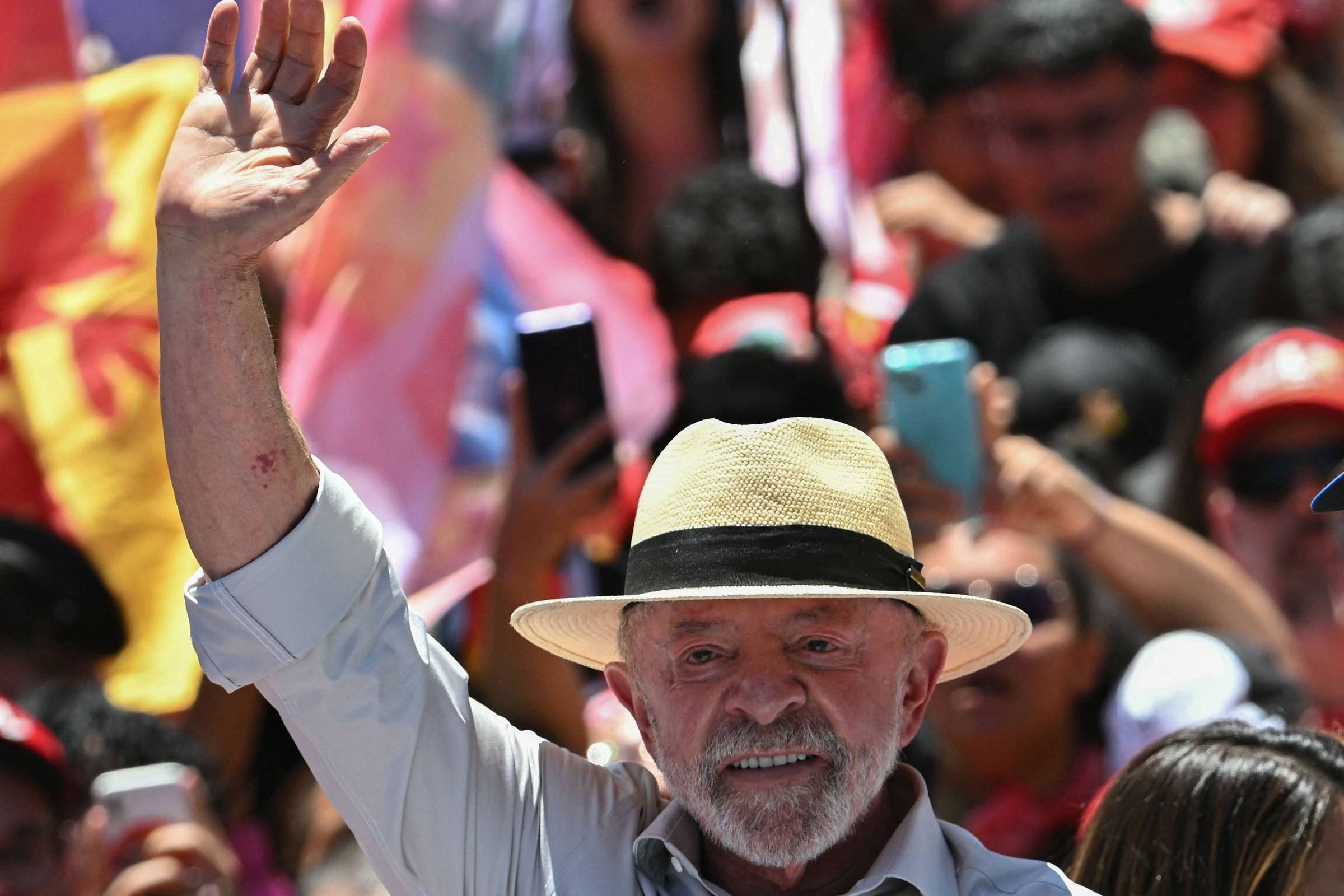 Homem de barba branca e chapéu de palha levanta a mão direita em cumprimento diante de multidão desfocada com bandeiras coloridas e pessoas usando celulares.