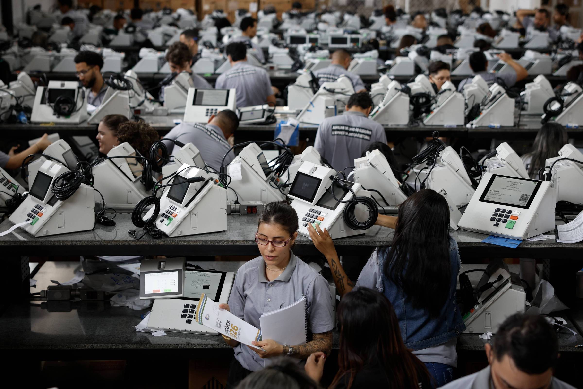 Diversos trabalhadores em uniforme cinza operam e inspecionam urnas eletrônicas alinhadas em longas mesas dentro de uma fábrica. O ambiente é amplo e cheio de equipamentos idênticos, com pessoas concentradas em tarefas manuais e de controle de qualidade.