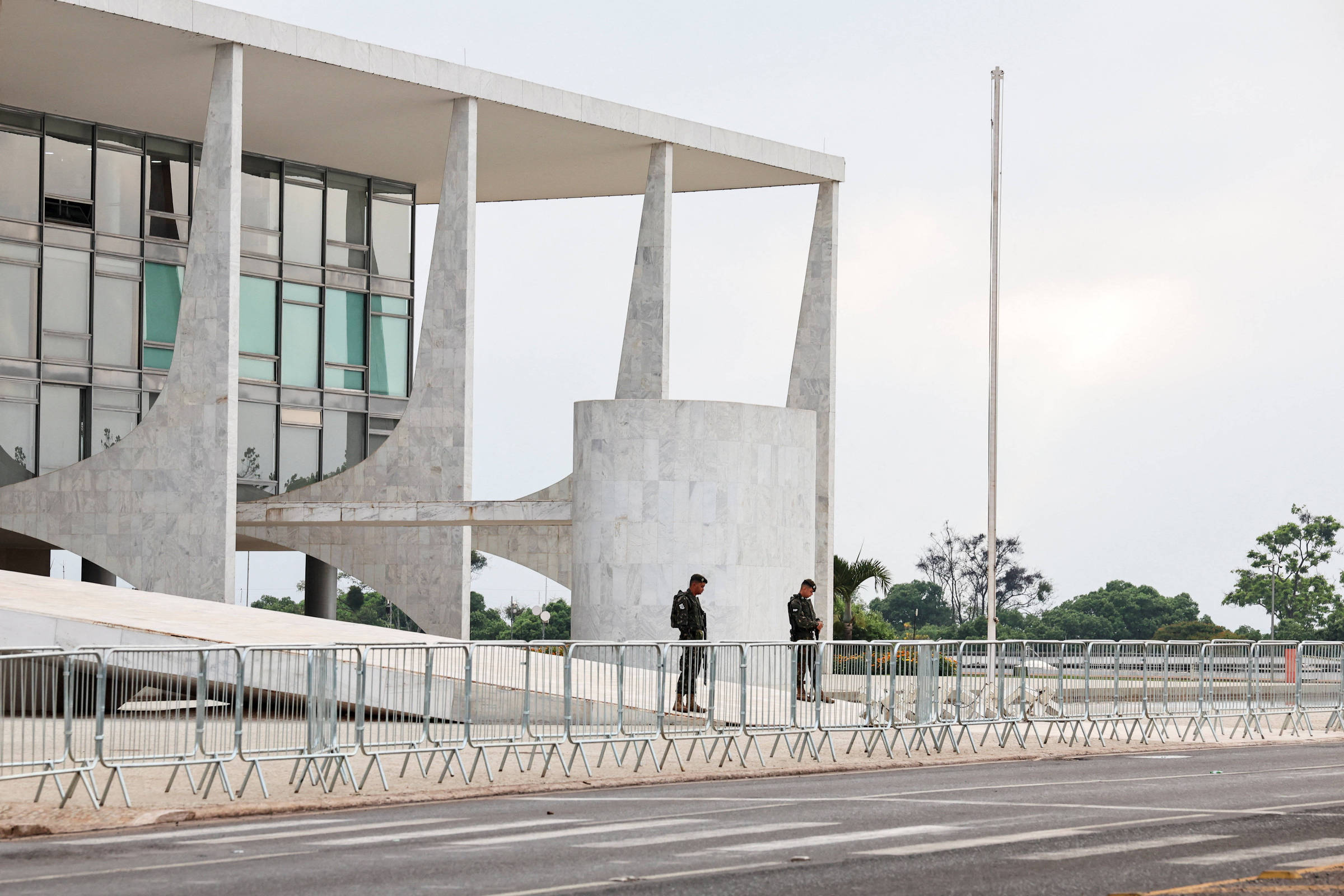 Dois seguranças estão posicionados próximos a um edifício público moderno com colunas de concreto e janelas de vidro. À frente, há várias barreiras metálicas alinhadas ao longo da calçada, e uma bandeira está hasteada em mastro vazio. O céu está claro e o ambiente parece tranquilo.