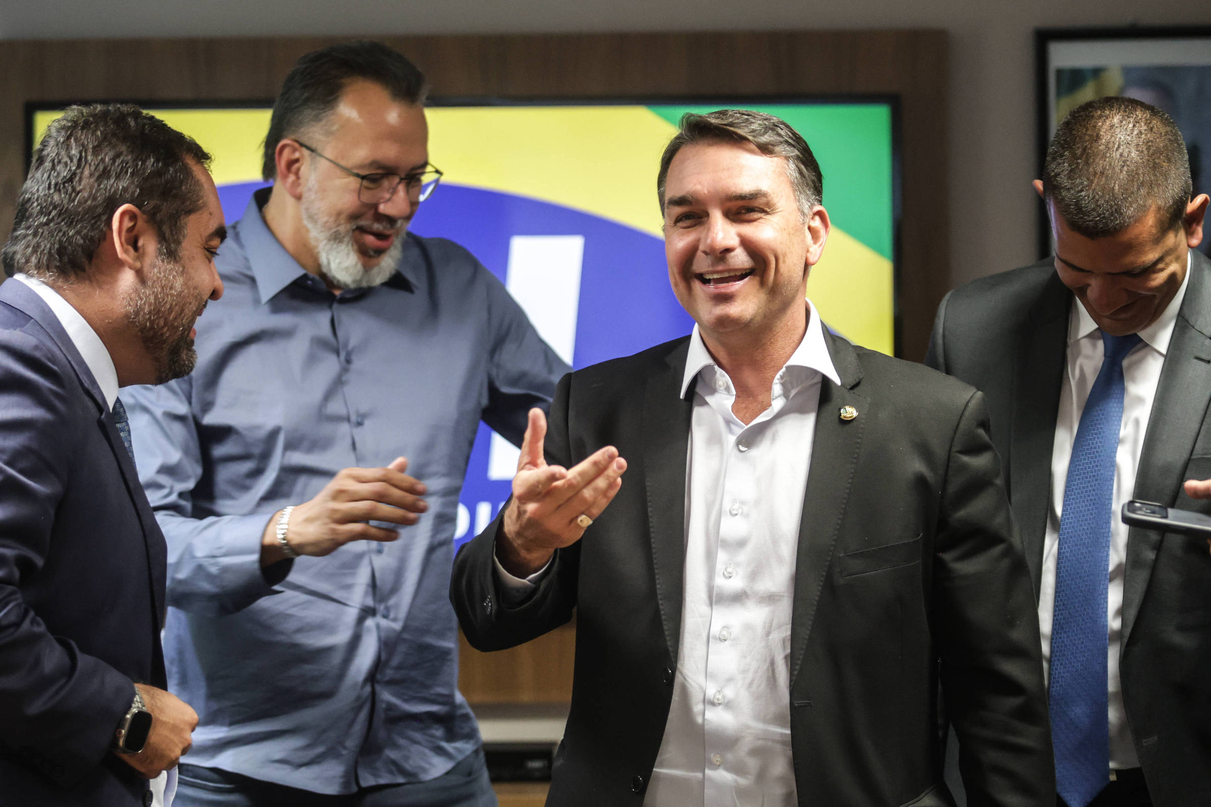 Quatro homens em trajes formais e casuais conversam e sorriem em ambiente interno. Ao fundo, tela exibe cores verde, amarela e azul, sugerindo bandeira do Brasil.