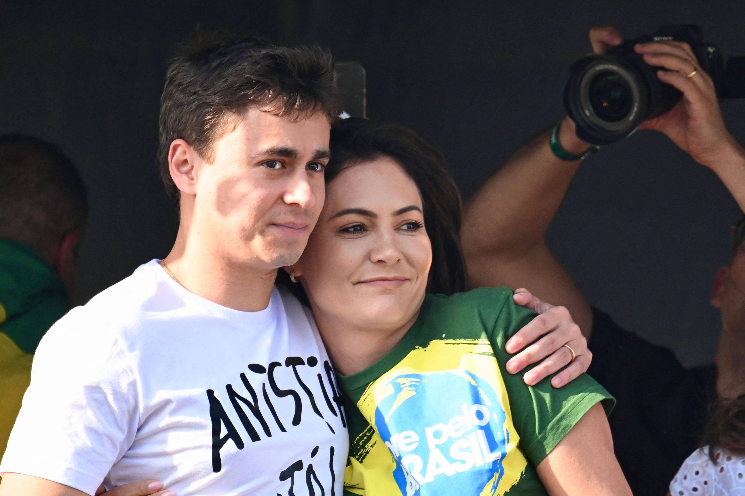 Homem e mulher abraçados, ele veste camiseta branca com texto preto e ela camiseta verde com estampa da bandeira do Brasil. Fundo escuro com pessoas e câmera fotográfica visível.